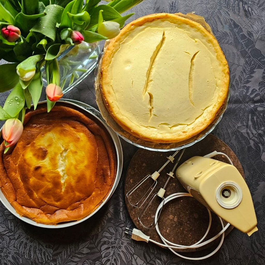 Zwei selbstgebackene Käsekuchen von oben fotografiert neben einem 50 Jahre alten Kalorik-Handmixer mit Kabel, dazwischen Tulpen auf gemusterter Tischdecke